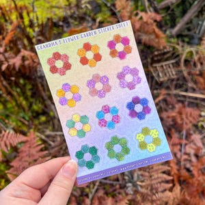 May include: A rectangular sticker sheet titled GRANDMA'S FLOWER GARDEN STICKER SHEET featuring twelve hexagonal flower motifs in various colors and patterns. The background has a soft gradient from yellow to purple, with text at the bottom.