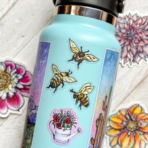May include: A light blue water bottle decorated with three individual bee stickers, a sticker of a white watering can filled with red and purple flowers, and a vertical rectangular sticker featuring a mountain landscape and a desert cactus scene.