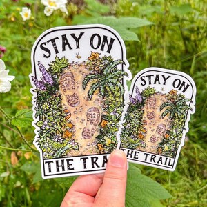 May include: Two die-cut stickers featuring an illustration of two muddy boot prints on a dirt path surrounded by green foliage, purple flowers, and yellow leaves. The text STAY ON THE TRAIL is printed in black capital letters at the top and bottom.
