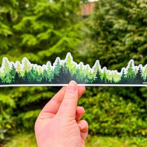 May include: A long, horizontal sticker featuring a watercolor-style illustration of a dense forest skyline. The design displays various shades of green, ranging from light lime to deep forest green, with a white border outlining the tree shapes.