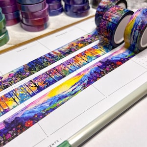 May include: Three rolls of decorative adhesive tape featuring a vibrant, colorful landscape illustration of mountains, trees, and flowers. The tape is unrolled across a white planner page with grid lines, showcasing a repeating scenic pattern.