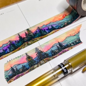 May include: Decorative washi tape featuring a watercolor landscape with mountains, trees, and a sunset in shades of pink, orange, and purple. A gold pen and a planner page with the days of the week are also visible.