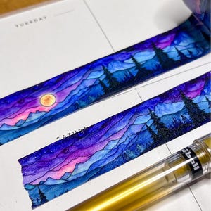 May include: Decorative washi tape featuring a vibrant watercolor-style landscape of layered blue and purple mountains, dark evergreen trees, and a glowing yellow moon against a starry night sky, applied to a white planner page with a gold pen.