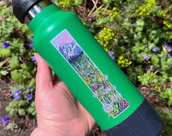 May include: A bright green Hydro Flask water bottle featuring a tall, rectangular sticker with a mountain landscape and a vibrant wildflower meadow. The bottle has a black screw-top lid with a carry loop and a black protective silicone boot base.