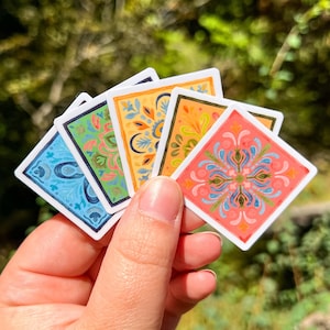 May include: A set of five square stickers arranged in a fan shape, each featuring a unique, colorful floral pattern. The designs include shades of blue, green, yellow, orange, and coral, all bordered by a thin white edge on a square base.