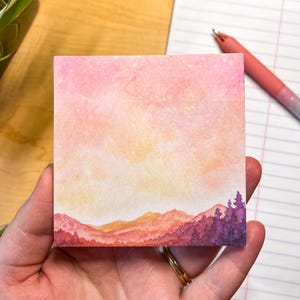 May include: A square sticky note featuring a watercolor landscape illustration. The design displays a gradient sky transitioning from soft pink to pale yellow above a range of orange mountains and a foreground of deep purple evergreen trees.
