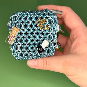 May include: A small, teal blue chainmail pouch with various charms attached, including a glass vial with dried flowers, a gold-colored charm, a black and white charm, and a small, iridescent bead.