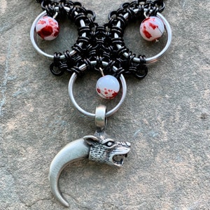 May include: A silver necklace with a wolf's head pendant and three white beads with red accents. The necklace is made of black chainmail.