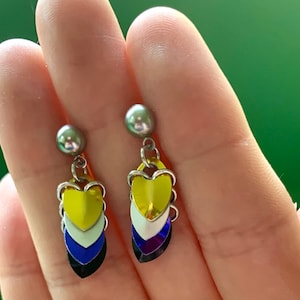Könnte beinhalten: Ein Paar Hängeohrringe mit einem mehrschichtigen Herzdesign. Die Herzen sind gelb, weiß, blau und schwarz. Die Ohrringe sind silberfarben und haben oben eine kleine silberne Perle.