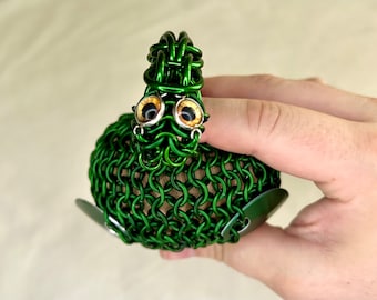 May include: A green metal sculpture of a dragon with large, round, brown eyes. The dragon is made of chainmail and has a long, thin tail.