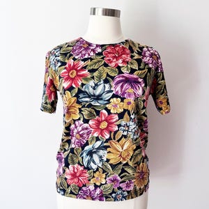 May include: A short-sleeved shirt with a vibrant floral pattern. The design features large, colorful flowers in shades of red, purple, yellow, and white against a dark navy blue background. The shirt is displayed on a mannequin.