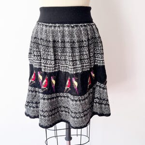 May include: A black and white patterned skirt with a black waistband and hem. The skirt features a repeating geometric design and a row of colorful bird motifs. The skirt is displayed on a mannequin.