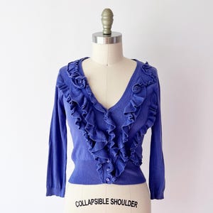 Op de afbeelding: Een periwinkle blauw vest met een V-hals en lange mouwen. De voorkant heeft een trapsgewijs ruchesontwerp. Het kledingstuk wordt getoond op een paspop met de tekst "COLLAPSIBLE SHOULDER" en het nummer "8".