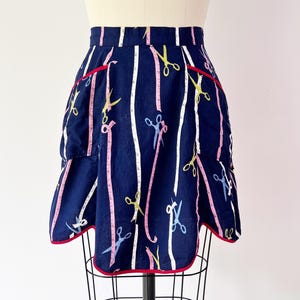 Może przedstawiać: Granatowy fartuch z czerwonym obszyciem i kieszeniami. Fartuch ma powtarzający się wzór białych taśm mierniczych, różowych taśm mierniczych i kolorowych nożyczek. Fartuch jest prezentowany na manekinie.