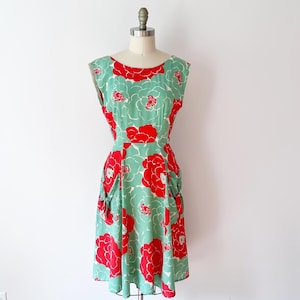 Może przedstawiać: Sukienka w stylu vintage na manekinie. Sukienka ma miętowo-zielone tło z dużymi czerwonymi wzorami kwiatowymi. Ma okrągły dekolt, kr&oacute;tkie rękawy i kieszenie. Sukienka jest do kolan.