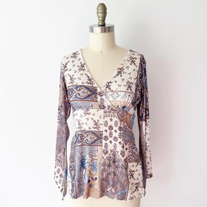 May include: A long-sleeved top with a V-neck, displayed on a mannequin. The top features a patchwork design with floral and paisley patterns in cream, blue, and brown tones. The sleeves are long and flared, and the fabric appears lightweight.