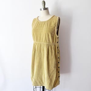 Op de afbeelding: Een mouwloze, mosterdgele corduroy jurk, gepresenteerd op een mannequin. De jurk heeft een ronde halslijn, een getailleerde taille en een rij donkere knopen aan de zijkant. De stof heeft een geribbelde textuur.