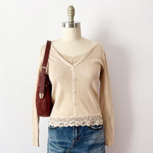 Puede incluir: Cárdigan beige acanalado con cuello en V y cierre de botones, combinado con una falda vaquera. El cárdigan presenta un ribete de encaje en el escote y el dobladillo. Un bolso de hombro de ante marrón oscuro con detalles plateados completa el conjunto.