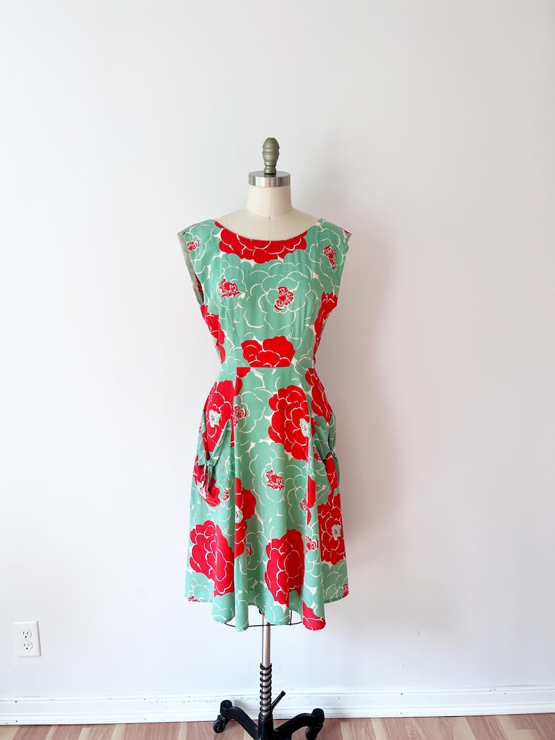 Może przedstawiać: Sukienka w stylu vintage na manekinie. Sukienka ma miętowo-zielone tło z dużymi czerwonymi wzorami kwiatowymi. Ma okrągły dekolt, kr&oacute;tkie rękawy i kieszenie. Sukienka jest do kolan.