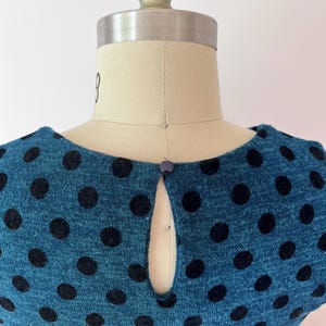 May include: Teal dress with large black polka dots. The dress has a keyhole neckline and a small button fastening. The fabric appears to be a knit material. The dress is displayed on a mannequin.
