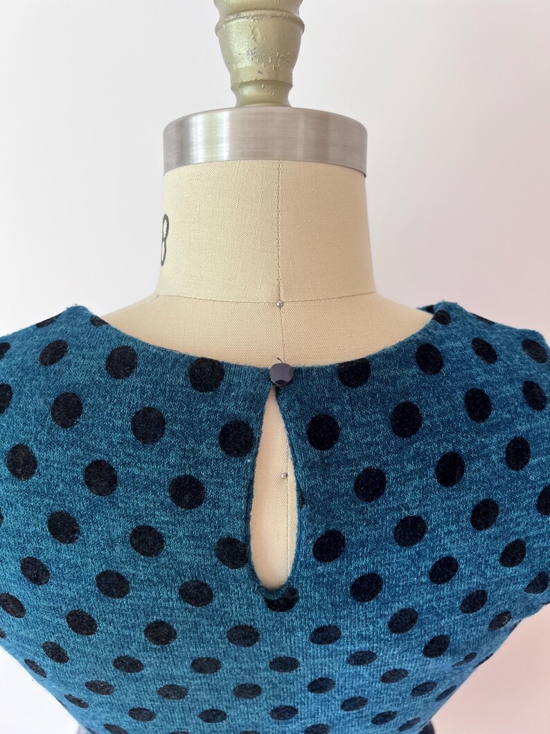 May include: Teal dress with large black polka dots. The dress has a keyhole neckline and a small button fastening. The fabric appears to be a knit material. The dress is displayed on a mannequin.