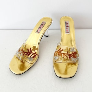 May include: A pair of gold-colored heeled sandals with clear straps and a floral design. The sandals have a square toe and a clear heel. The brand name "s.t.e.f.a.n.i" is printed on the insole.