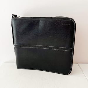 May include: Black leather messenger bag with a shoulder strap. The bag features white stitching details and a horizontal band across the front. The zipper runs along the top edge. The brand name is visible on the top right corner.