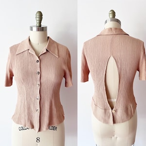 May include: A light pink short-sleeved blouse with a collared neckline and button-down front. The back of the blouse features a keyhole design. The blouse is displayed on a mannequin.