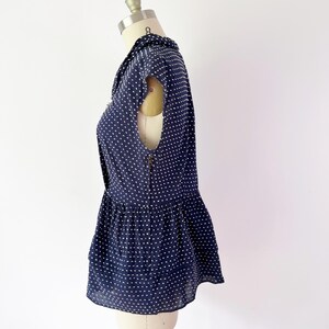 May include: Navy blue sleeveless blouse with white polka dots. The blouse features a collared neckline, a peplum hem, and a zipper closure down the back. The garment is displayed on a mannequin.