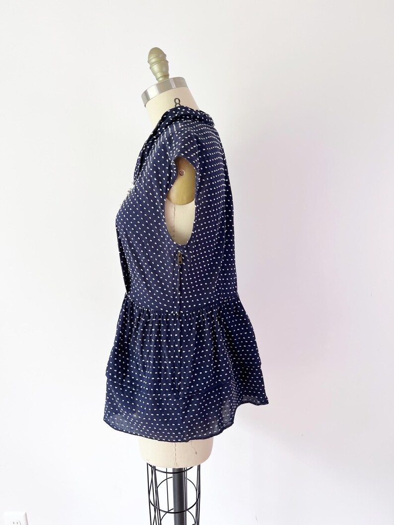 May include: Navy blue sleeveless blouse with white polka dots. The blouse features a collared neckline, a peplum hem, and a zipper closure down the back. The garment is displayed on a mannequin.
