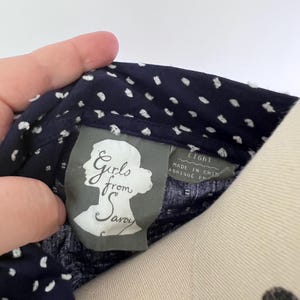 May include: Close-up of a navy blue fabric with white polka dots. The tag reads "Girls from Savoy" and "EIGHT". The fabric is held by a hand. The tag is gray with a silhouette of a woman's head.