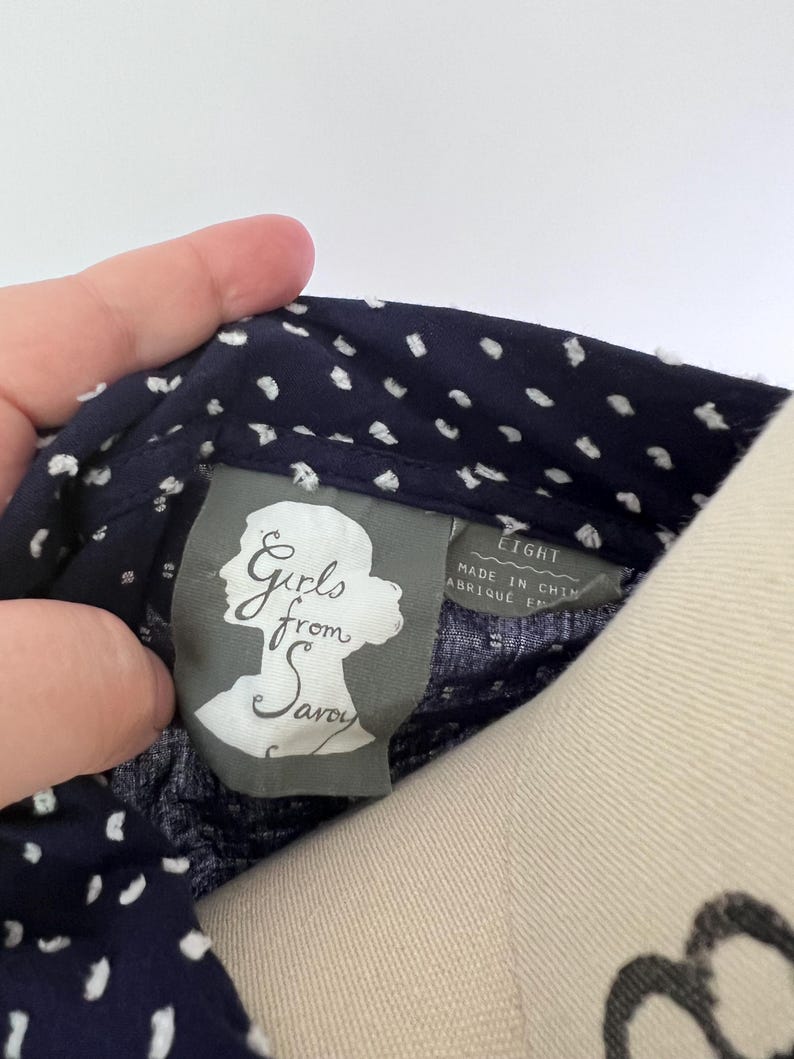 May include: Close-up of a navy blue fabric with white polka dots. The tag reads "Girls from Savoy" and "EIGHT". The fabric is held by a hand. The tag is gray with a silhouette of a woman's head.