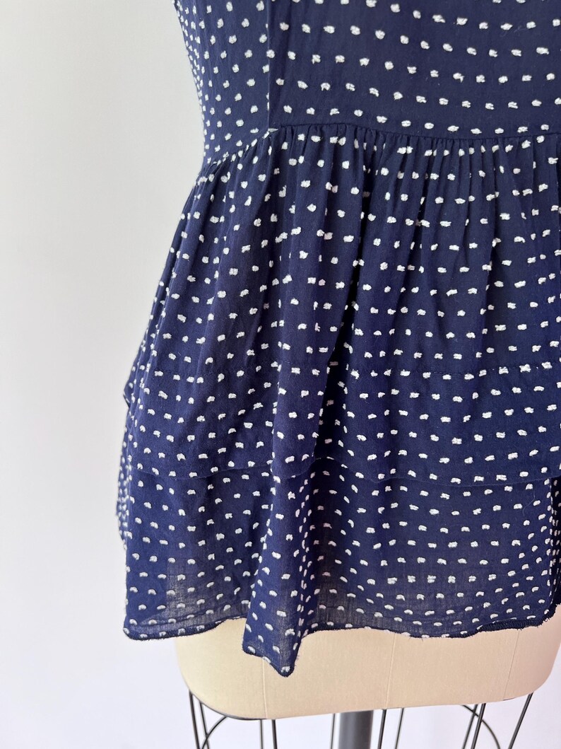 May include: Navy blue top with white polka dots. The top features a tiered, peplum-style design. The fabric appears lightweight and textured, with a subtle crinkled effect. The garment is displayed on a mannequin.