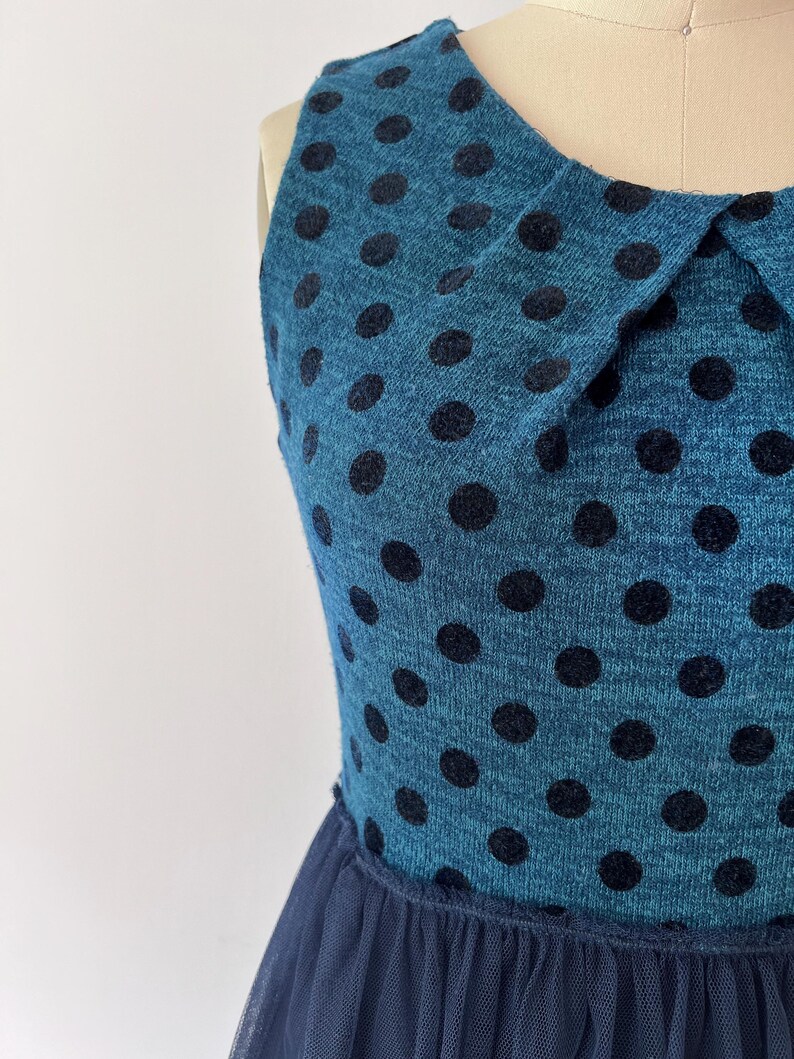 May include: A teal dress with black polka dots and a navy blue tulle skirt. The sleeveless dress has a Peter Pan collar. The fabric has a textured appearance, and the polka dots are evenly spaced across the bodice.