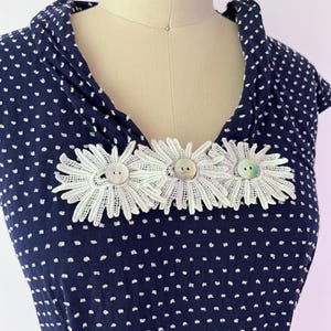 May include: Navy blue dress with white polka dots, featuring a cowl neckline. The dress is adorned with three white lace daisy embellishments, each centered with a button. The dress is displayed on a mannequin.