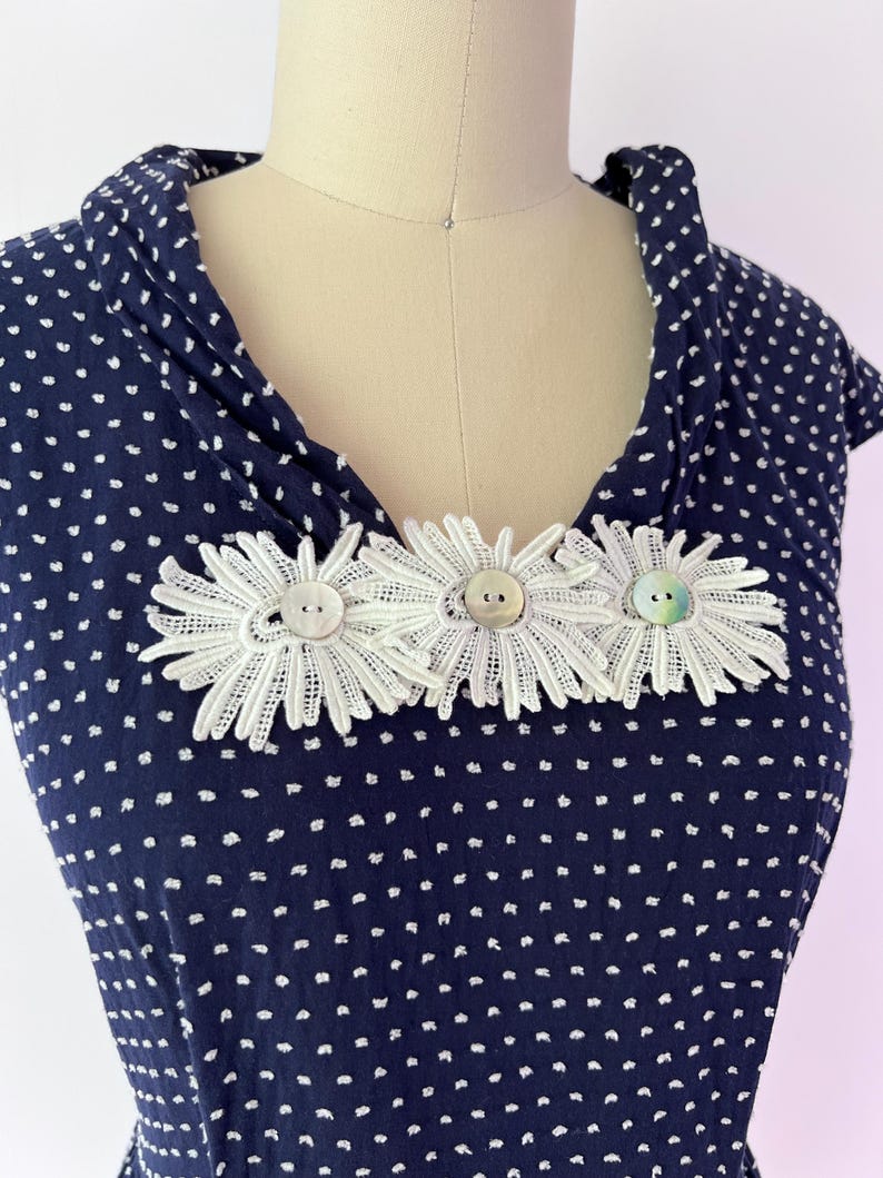 May include: Navy blue dress with white polka dots, featuring a cowl neckline. The dress is adorned with three white lace daisy embellishments, each centered with a button. The dress is displayed on a mannequin.