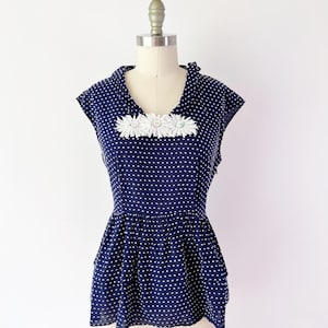 May include: Navy blue polka dot top with white daisy embellishments. The top features a peplum hem and is displayed on a mannequin. The top has a V-neckline and short sleeves. The fabric is lightweight and semi-sheer.