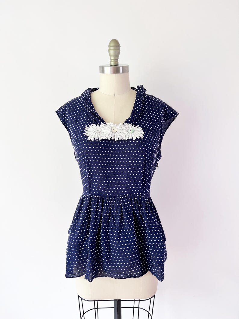 May include: Navy blue polka dot top with white daisy embellishments. The top features a peplum hem and is displayed on a mannequin. The top has a V-neckline and short sleeves. The fabric is lightweight and semi-sheer.