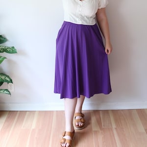May include: A solid purple midi skirt with a flared silhouette and an elasticized waistband, paired with a cream-colored short-sleeved top featuring a button-down front and brown leather sandals with two wide straps and a buckle closure.