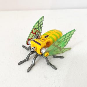 May include: A yellow and green toy insect with transparent green wings. The insect has a yellow body with green stripes, gray legs, and a silver-colored head. The wings are a transparent green with a grid-like pattern.