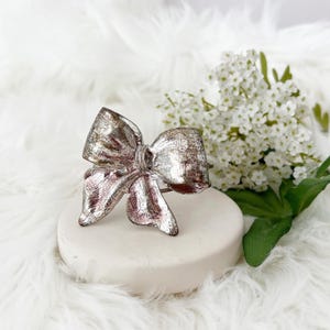 Puede incluir: Un anillo plateado con forma de lazo y acabado texturizado. El anillo está colocado sobre un pedestal redondo blanco, con flores blancas y vegetación en el fondo. El diseño del lazo es detallado y llamativo, adecuado para una ocasión especial.