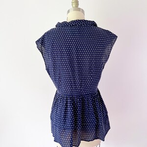 May include: Navy blue blouse with white polka dots. The blouse features a collared neckline, short sleeves, and a gathered waist. The fabric appears lightweight and semi-sheer, with a slight sheen. The blouse is displayed on a mannequin.
