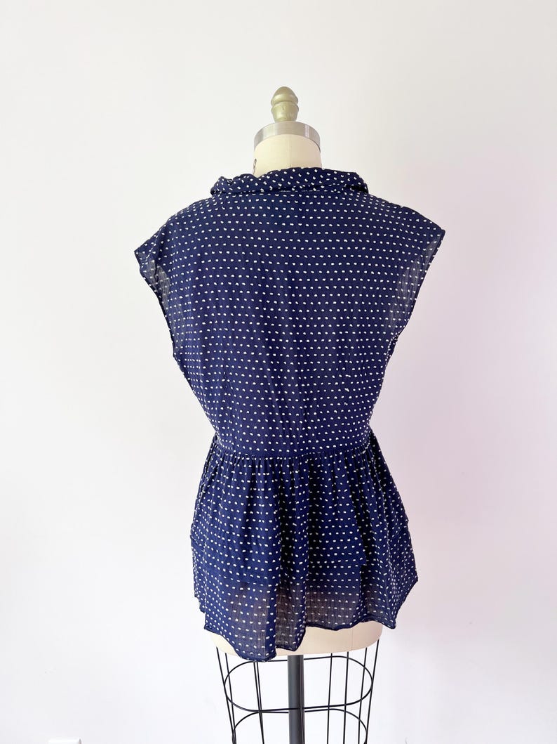 May include: Navy blue blouse with white polka dots. The blouse features a collared neckline, short sleeves, and a gathered waist. The fabric appears lightweight and semi-sheer, with a slight sheen. The blouse is displayed on a mannequin.