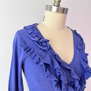 Op de afbeelding: Een periwinkle blauw vest met een V-hals en lange mouwen. Het vest heeft ruches langs de halslijn en voorkant, met een klein rozetdetail aan de bovenkant. Het kledingstuk wordt op een mannequin getoond.