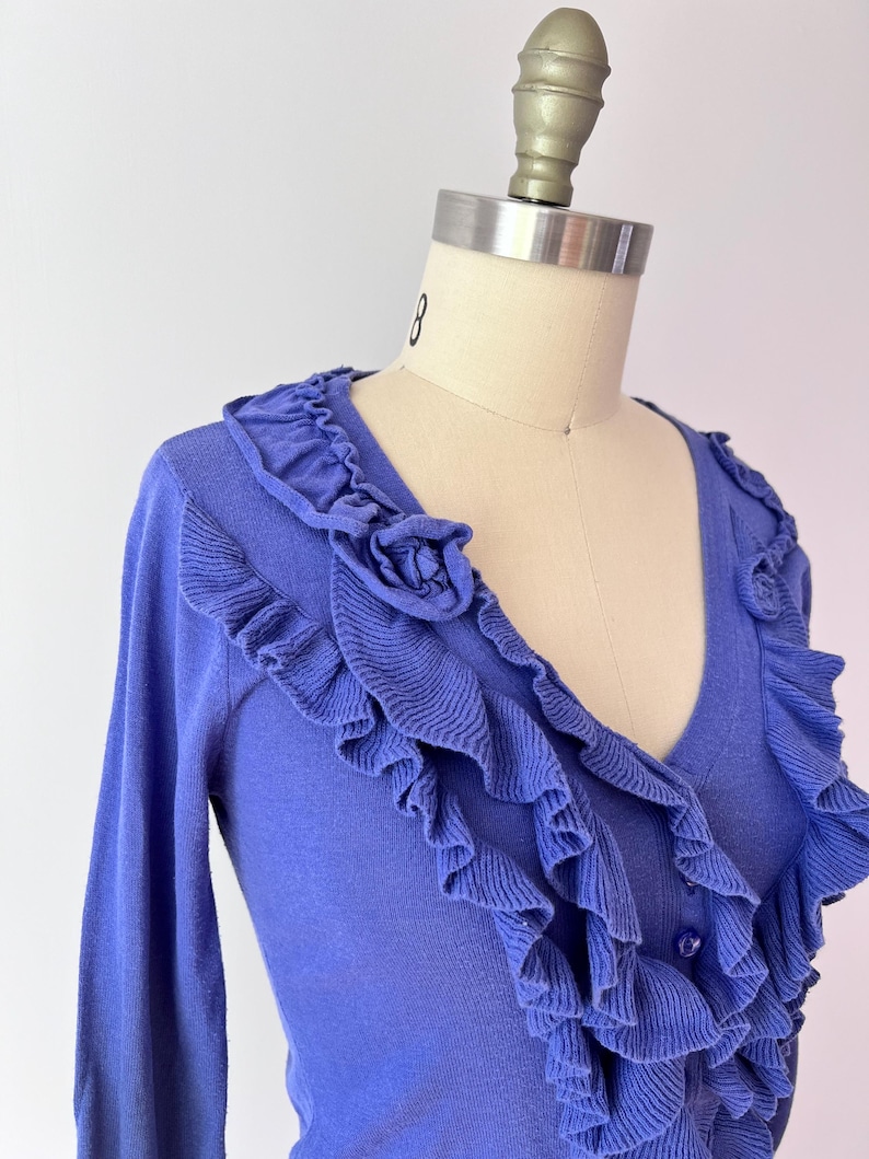 Op de afbeelding: Een periwinkle blauw vest met een V-hals en lange mouwen. Het vest heeft ruches langs de halslijn en voorkant, met een klein rozetdetail aan de bovenkant. Het kledingstuk wordt op een mannequin getoond.