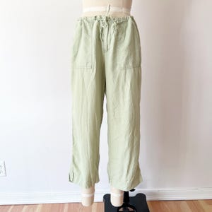 Peut inclure: Pantalon en lin vert clair avec une taille élastique et deux poches avant. Le pantalon a une coupe décontractée et une longueur raccourcie, avec une petite fente à l'ourlet. Le pantalon est présenté sur un mannequin.