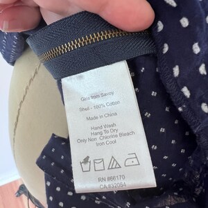 May include: Close-up of a navy blue fabric with white polka dots. The fabric is attached to a zipper. A tag reads "Girls from Savoy" and lists care instructions: hand wash, hang to dry, non-chlorine bleach, and iron cool. RN #66170, CA #32054.
