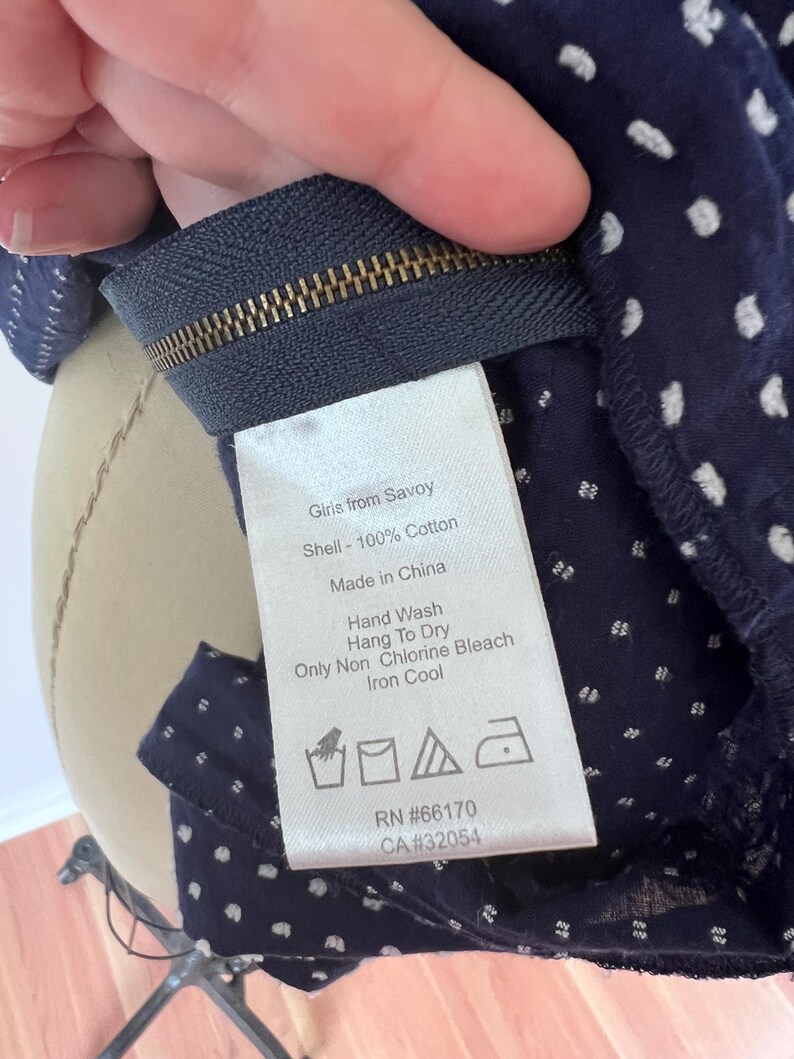 May include: Close-up of a navy blue fabric with white polka dots. The fabric is attached to a zipper. A tag reads "Girls from Savoy" and lists care instructions: hand wash, hang to dry, non-chlorine bleach, and iron cool. RN #66170, CA #32054.