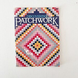 May include: A vintage book titled "Discovering Patchwork" with a colorful patchwork design on the cover. The cover features a geometric pattern in shades of red, pink, yellow, gray, and black. The book is set against a white background.