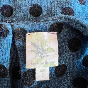 May include: Close-up of a blue knit fabric with large black polka dots. A tag is visible, with a bird illustration and text that reads "Made in San Francisco" and "USA S". The fabric appears to be a top or dress.